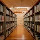 Estantes de biblioteca repletos de libros se alinean a ambos lados de un amplio pasillo de madera. Los estantes son altos y están abarrotados de libros diversos, con el lomo hacia afuera. El suelo es de madera pulida y la iluminación es cálida, provenient