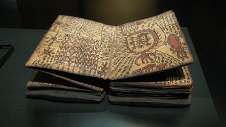 Manuscrito antiguo con símbolos y dibujos misteriosos. El libro abierto presenta páginas gruesas y manchadas, cubiertas de intrincados patrones, cuadrículas y glifos inusuales que parecen dibujados a mano. La página izquierda presenta formas geométricas y