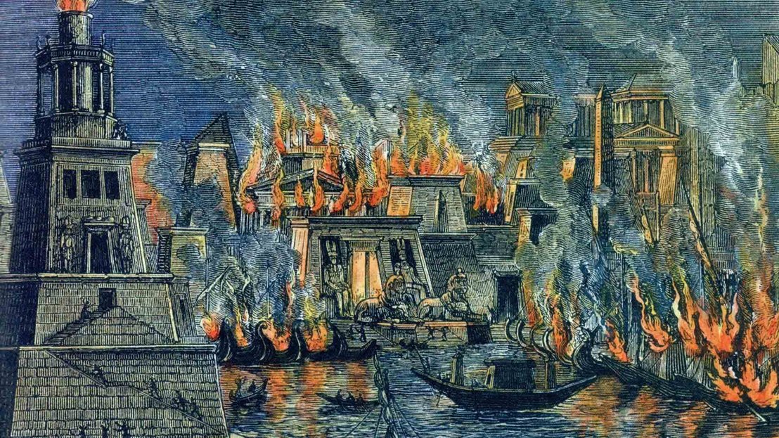 Ilustración histórica de un incendio urbano, probablemente el de la Biblioteca de Alejandría. La imagen muestra edificios de estilo griego y egipcio clásico envueltos en llamas, con un fuego naranja brillante y una densa humareda elevándose hacia el cielo