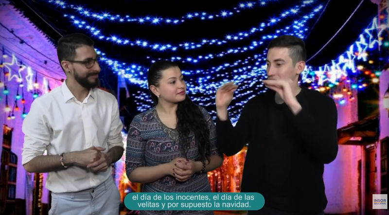Tres personas posan frente a un fondo festivo con luces azules y blancas en forma de estrella que decoran una calle. El hombre de la izquierda lleva gafas y una camisa blanca; la mujer del centro tiene el pelo largo y oscuro y viste un vestido estampado; y el hombre de la derecha viste de negro y parece estar hablando o haciendo señas con las manos levantadas. En la parte inferior de la imagen, aparece un subtítulo en español que dice: «El día de los inocentes, el día de las velitas y, por supuesto, la Navidad». En la esquina inferior derecha, hay un pequeño logotipo que dice «INSOR EDUCATIVO». La escena sugiere una charla sobre las tradiciones navideñas, probablemente relacionadas con la Navidad y otras celebraciones.
